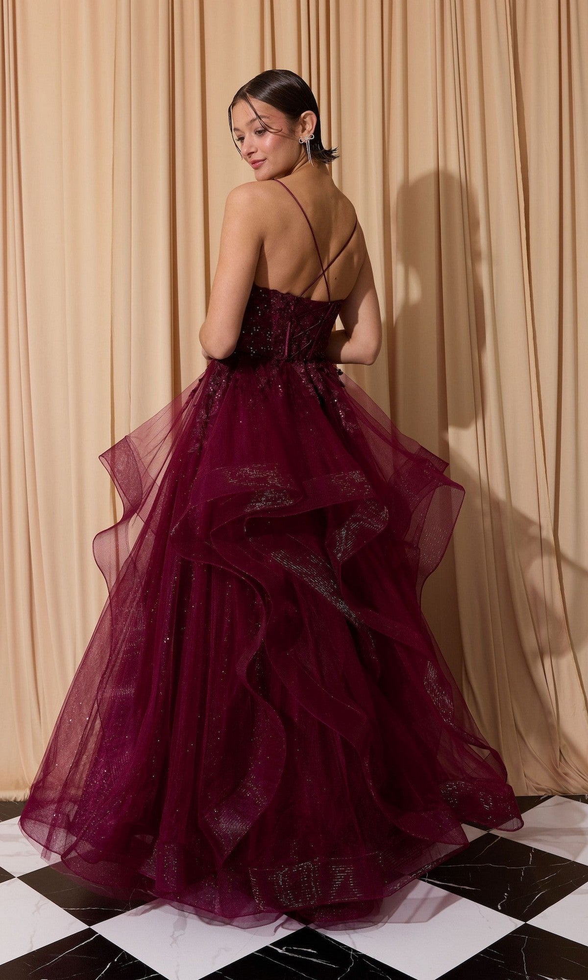 A woman wearing the Long Prom Dress: Eureka EK167, featuring a burgundy layered tulle A-line skirt and spaghetti straps, stands on a black-and-white tiled floor before beige curtains, smiling as she faces slightly away.